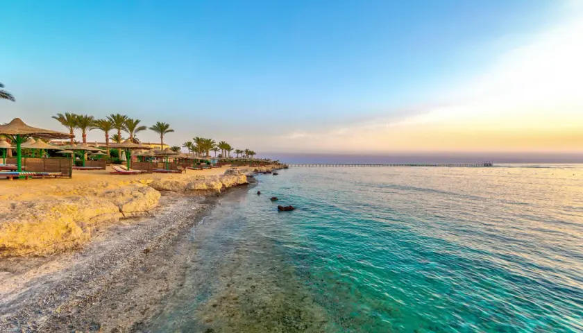 Piękna pogoda w Marsa Alam: piaszczysta plaża z palmami, leżakami i parasolami, turkusowe morze i długie molo na horyzoncie.