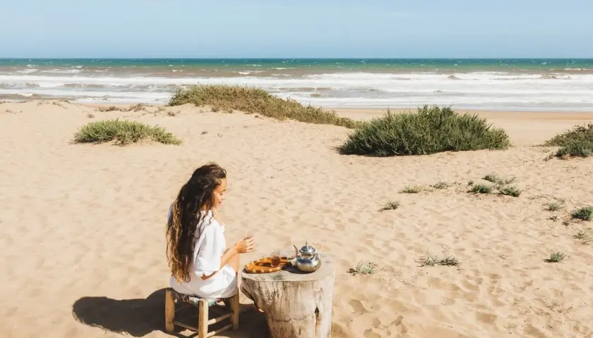Kobieta pije herbatę na piaszczystej plaży. W tle widać turkusowe morze, które przypomina o pięknie Tunezji.