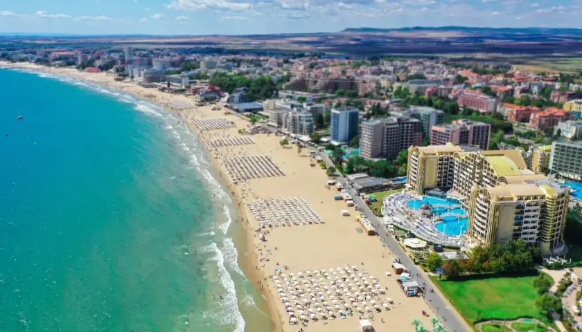 Słoneczna riwiera bułgarska z szeroką, piaszczystą plażą pełną leżaków i parasoli, turkusowym morzem i hotelami w tle.