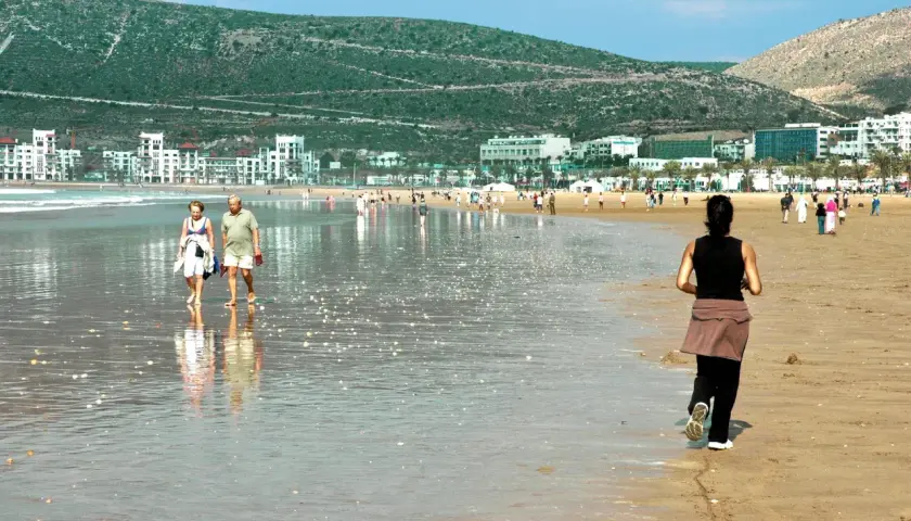Spacerem po plaży w Maroku, w tle białe budynki i zielone wzgórza. Idealne wakacje.