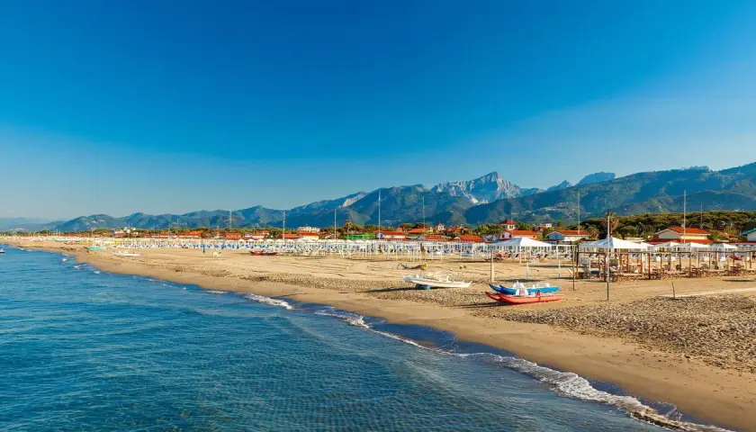 Słoneczna plaża w Toskanii, z rzędami białych parasoli i leżaków, w tle góry i błękitne morze.