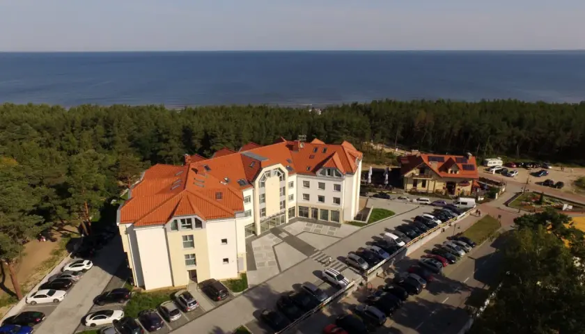 Hotel blisko morza, idealny na wypad z Śląska. Widać parking pełen aut, las i błękitną wodę.