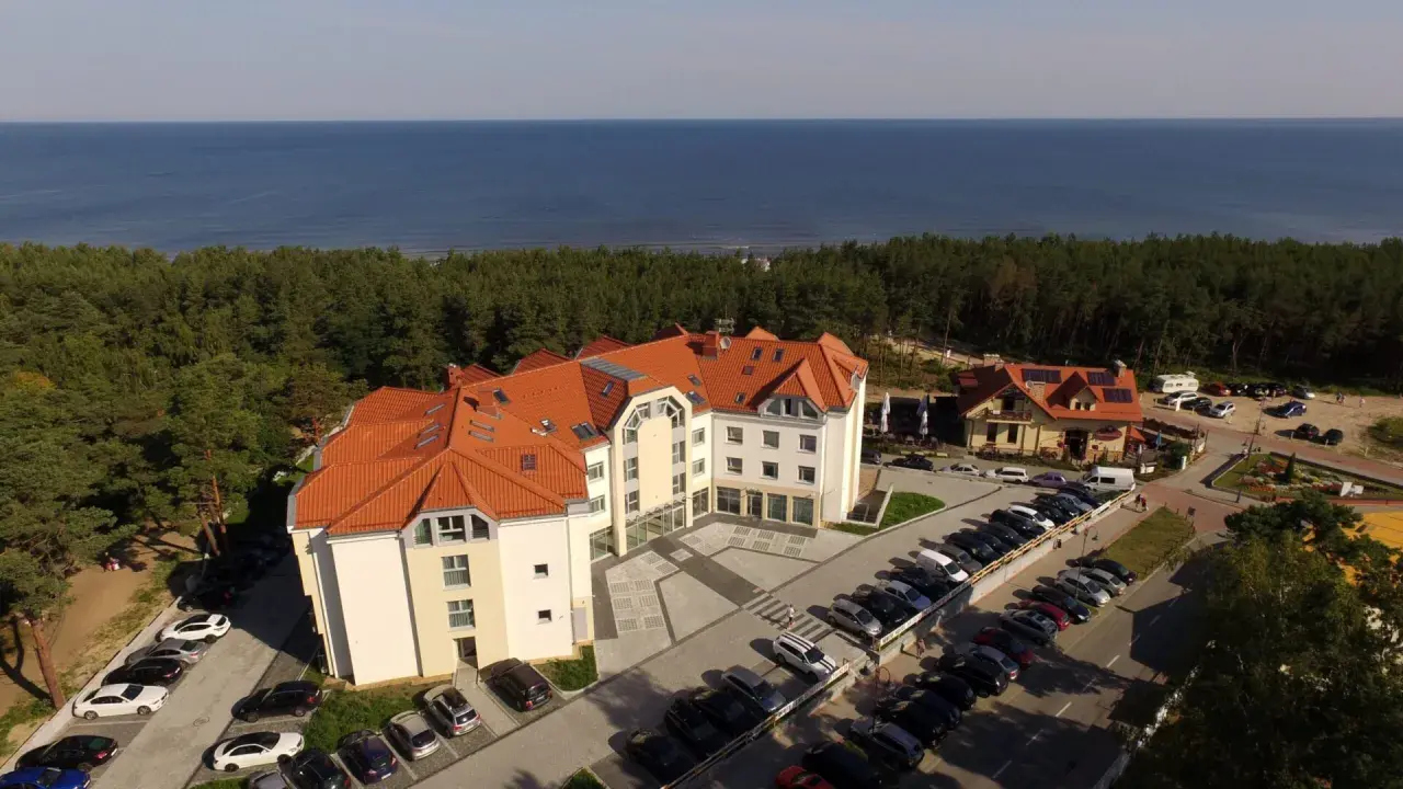 Hotel blisko morza, idealny na wypad z Śląska. Widać parking pełen aut, las i błękitną wodę.