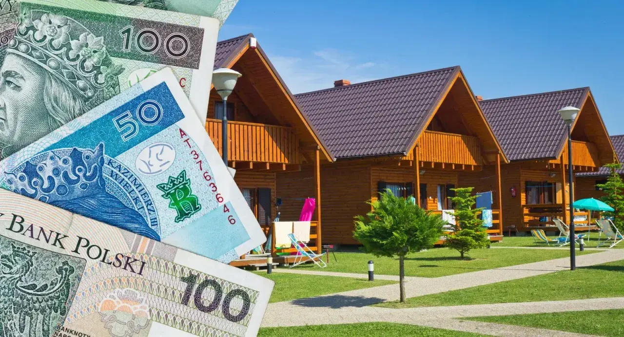 Polskie banknoty na tle drewnianych domków. Szukasz sposobu, jak znaleźć tani nocleg?